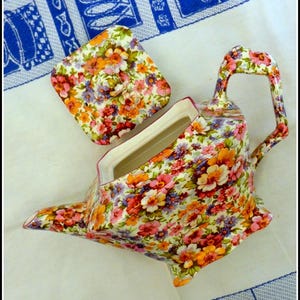 Pode incluir: Um bule de cerâmica com tampa quadrada, apresentando um padrão floral em tons de rosa, laranja, amarelo e roxo. O bule possui uma alça curva, um bico pontiagudo e um interior na cor creme, apresentado a partir de um ângulo superior.
