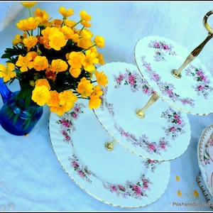 May include: A three-tiered cake stand with a floral design and gold detailing. The plates are white with pink and purple flowers. A blue vase holds yellow flowers. A teacup and saucer are visible.