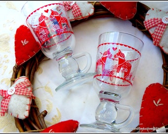 Könnte beinhalten: Zwei klare Glasbecher mit roten und weißen Motiven von stilisierten Pferden. Die Becher haben einen Henkel und sind mit einem roten gewellten Rand verziert. Die Becher stehen auf einem braunen Weidenkranz mit roten Filzherzen und weißen Filzherzen mit roten und weißen Karobändern.
