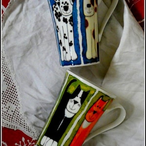 Peut inclure: Deux mugs en céramique avec des illustrations d'animaux. L'un représente un dalmatien et un chien marron et blanc sur fond bleu. L'autre présente un chat noir et blanc et un chat orange sur fond vert. Les deux mugs ont des anses blanches.