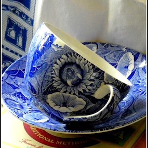 Pode incluir: Um conjunto de chávena e pires azul e branco com um design floral de girassol. A chávena está inclinada, revelando o seu interior branco. O conjunto é colocado numa superfície com o texto: "Traditional Methods Famous flavorful taste SINCE 1889".