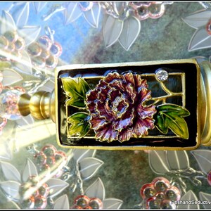 May include: A gold-toned bottle stopper with a black and green enamel design featuring a purple flower and a clear rhinestone.