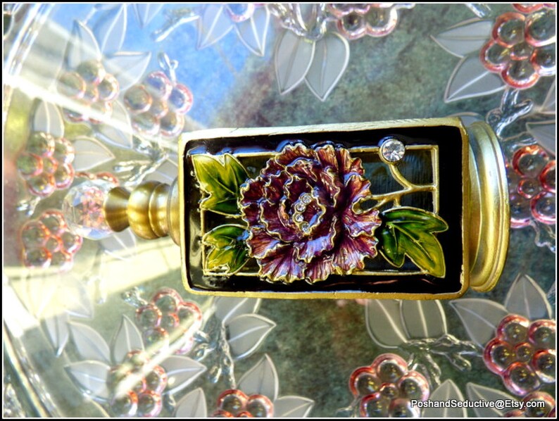 May include: A gold-toned bottle stopper with a black and green enamel design featuring a purple flower and a clear rhinestone.