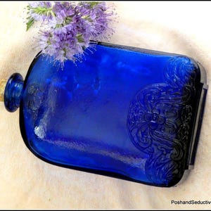 Peut inclure: Une bouteille en verre bleu cobalt avec un bouchon en liège et un motif floral en relief. La bouteille est posée sur une surface en tissu blanc.