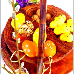 May include: A wooden tiered serving tray with a handle, featuring a decorative design. The tray is filled with Easter eggs, nuts, and small yellow chicks. A sprig of pussy willow branches is in the foreground.
