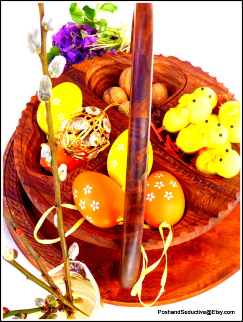 May include: A wooden tiered serving tray with a handle, featuring a decorative design. The tray is filled with Easter eggs, nuts, and small yellow chicks. A sprig of pussy willow branches is in the foreground.