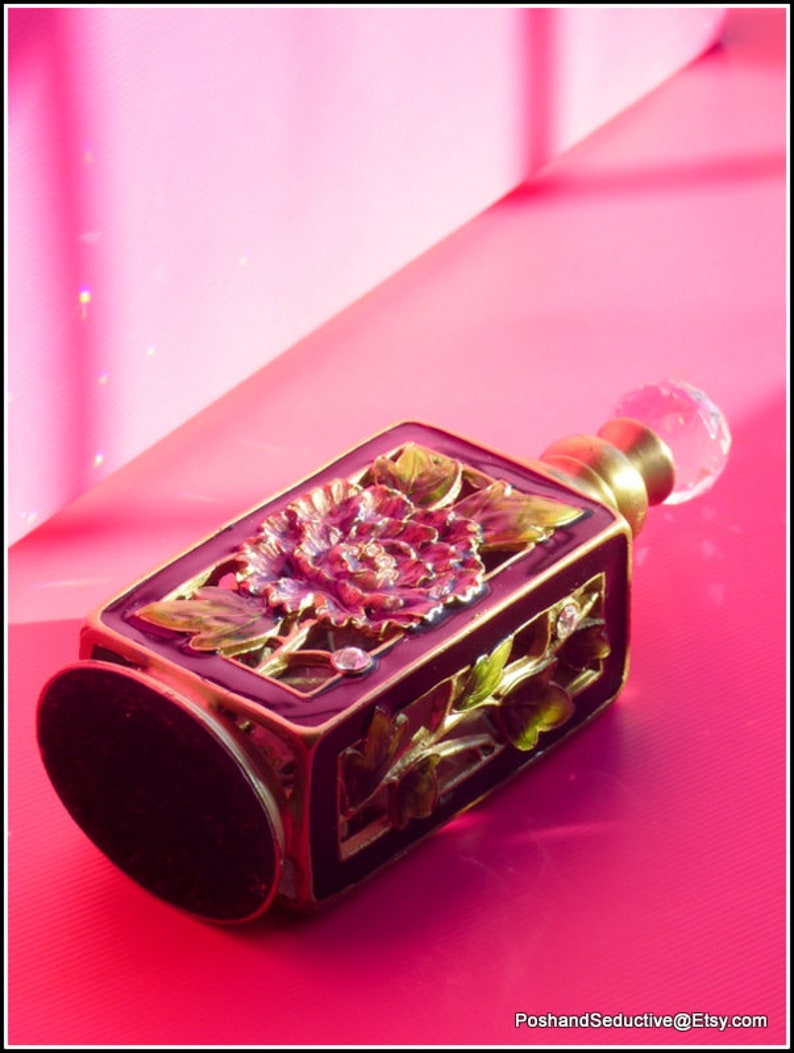 May include: A dark purple glass perfume bottle with a gold floral design and a clear glass stopper. The bottle is sitting on a pink surface.