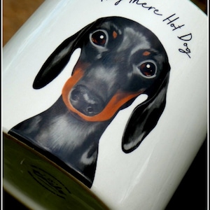 Könnte beinhalten: Weiße Keramiktasse mit einer Dackel-Illustration und dem Text "Hey there Hot Dog". Der Hund hat schwarzes Fell, braune Augen und eine orangefarbene Schnauze. Die Tasse hat einen grünen Henkel und eine glänzende Oberfläche.