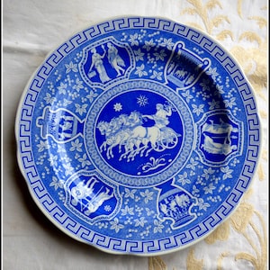 May include: A blue and white ceramic plate featuring a Greek key border and a central image of a chariot drawn by four horses. The plate is decorated with floral motifs and other scenes from Greek mythology.