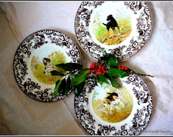 May include: Three brown and white ceramic plates with a floral design and a hunting dog illustration in the center. The plates are on a white tablecloth with a sprig of green leaves and red berries.