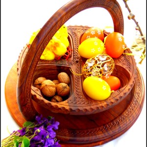May include: A wooden serving tray with four compartments, featuring a carved design. The tray holds yellow chicks, decorated Easter eggs, walnuts, and a crystal ornament. The tray is on a wooden base with a handle.