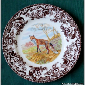 May include: A white ceramic dinner plate with a brown floral border and a painted image of a red fox in a woodland setting.