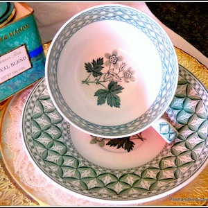 May include: A white teacup and saucer with a green and white floral pattern. The saucer has a geometric design with green and white accents. The teacup has a floral design with a green stem and white flowers. The teacup and saucer are on a gold and white tablecloth.