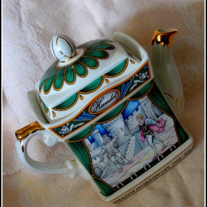 Op de afbeelding: Een witte keramische theepot met deksel, handvat en tuit, met een scène uit Hamlet. De theepot heeft gouden accenten en groene en blauwe decoratieve elementen. De tekst "Hamlet" en "William Shakespeare 1564-1616" zijn zichtbaar.