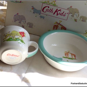 Op de afbeelding: Een witte keramische kinderset met een kom en een beker met een turquoise rand. De kom en de beker hebben een kleurrijk dierenprintontwerp met giraffen, tijgers en leeuwen. De beker heeft twee handvatten en de tekst "Cath Kidston" staat op de bodem.