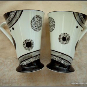 May include: Two black and white ceramic mugs with a geometric pattern. The mugs have a wide base and a narrow top.