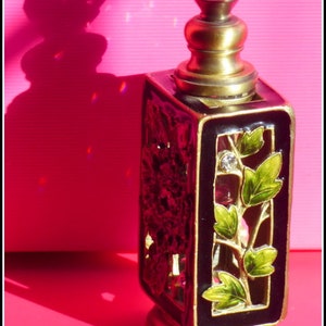 May include: A black and gold perfume bottle with a clear glass stopper. The bottle has a decorative design with green leaves and a floral pattern.