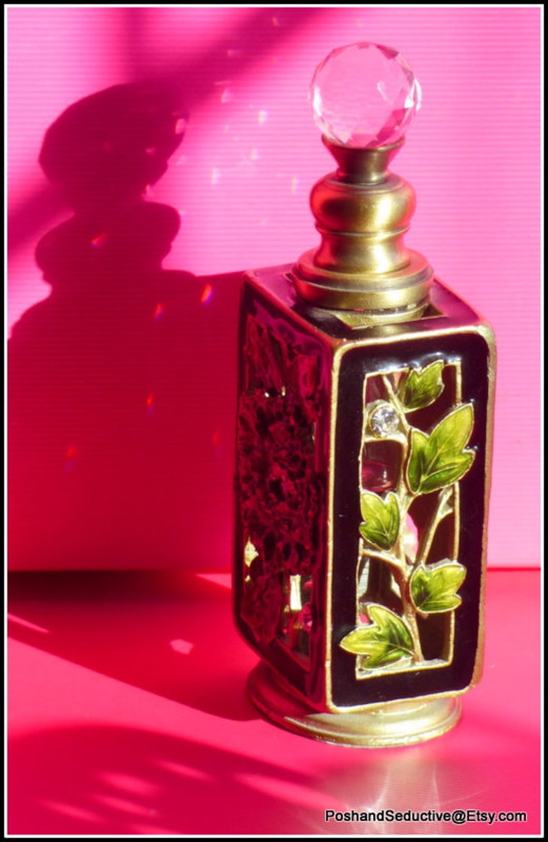May include: A black and gold perfume bottle with a clear glass stopper. The bottle has a decorative design with green leaves and a floral pattern.