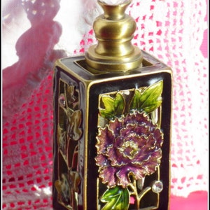 May include: A black and gold perfume bottle with a crystal stopper. The bottle is decorated with a floral design featuring a purple flower and green leaves.