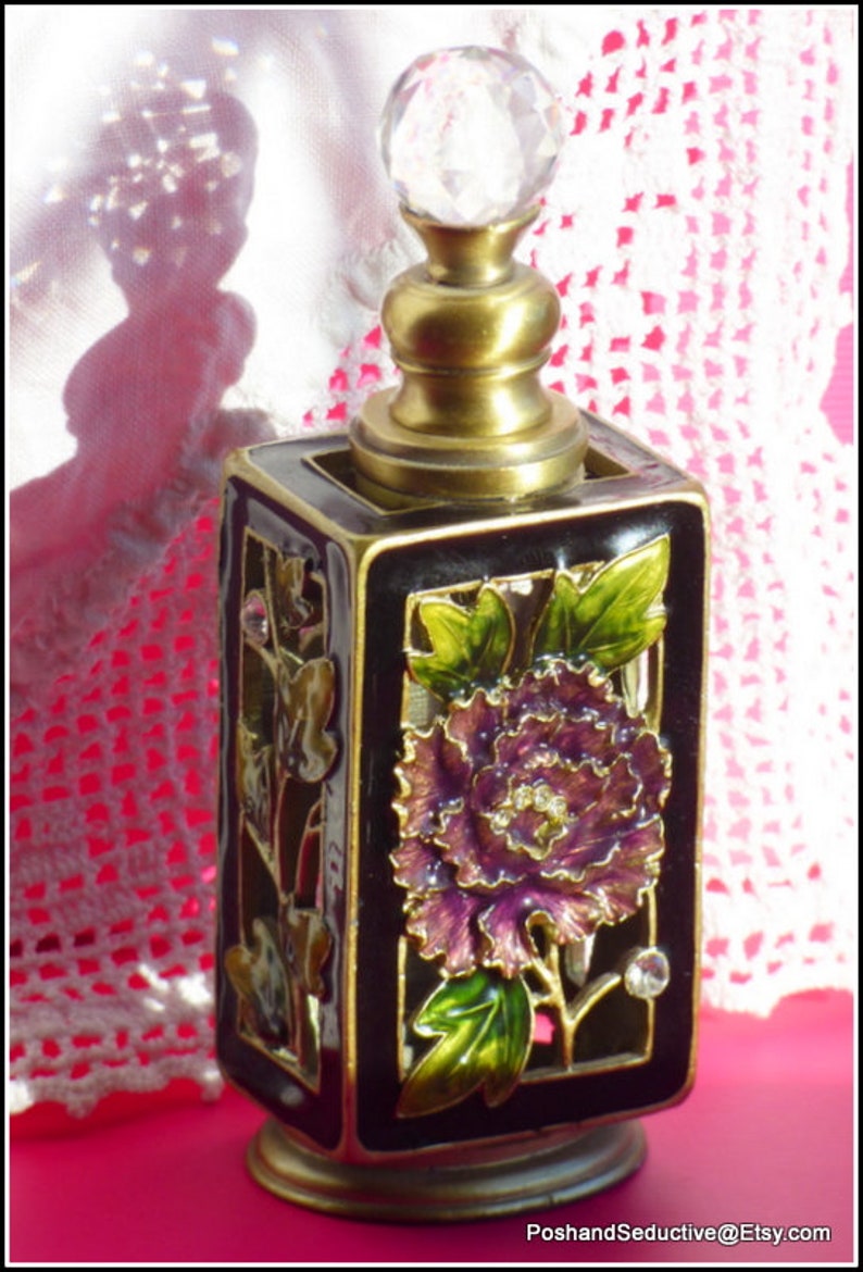 May include: A black and gold perfume bottle with a crystal stopper. The bottle is decorated with a floral design featuring a purple flower and green leaves.