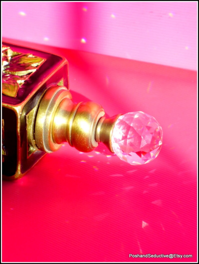 May include: A black and gold bottle stopper with a crystal-like knob. The stopper is on a pink surface.