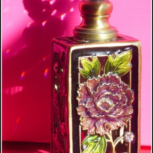 May include: A gold and black perfume bottle with a clear glass stopper. The bottle is decorated with a purple flower and green leaves.