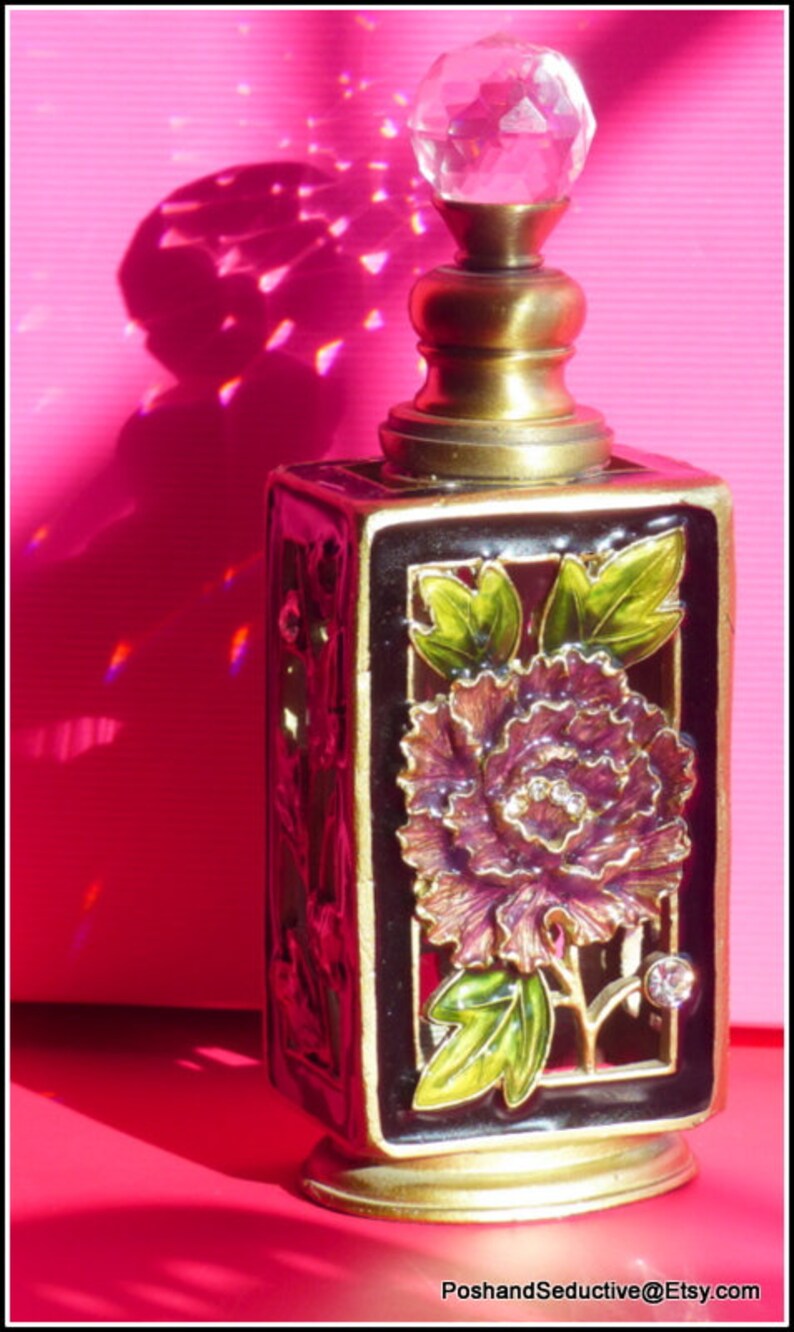 May include: A gold and black perfume bottle with a clear glass stopper. The bottle is decorated with a purple flower and green leaves.