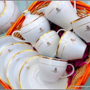 May include: A set of white teacups and saucers with gold trim and the letters "BR" on each piece. The cups and saucers are arranged in a wicker basket with a pink and orange floral pattern.