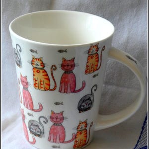 Könnte beinhalten: Ein weißer Keramikbecher mit einem wiederkehrenden Muster aus bunten Cartoon-Katzen in Orange-, Rosa- und Grautönen, durchsetzt mit kleinen grauen Fischillustrationen. Der Becher hat einen geschwungenen Henkel und eine glatte, glänzende Oberfläche.