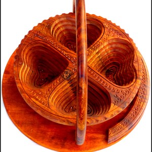 May include: A wooden fruit bowl with a handle, carved in a heart shape. The bowl is made of multiple layers of wood, and the handle is attached to the top of the bowl. The bowl is a rich reddish-brown color.