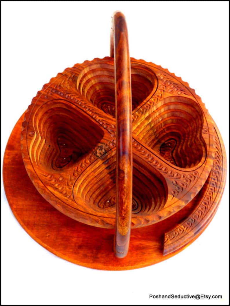 May include: A wooden fruit bowl with a handle, carved in a heart shape. The bowl is made of multiple layers of wood, and the handle is attached to the top of the bowl. The bowl is a rich reddish-brown color.
