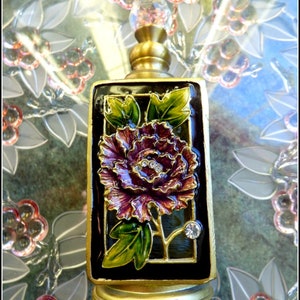 May include: A gold-toned perfume bottle with a black enamel frame and a floral design. The bottle features a large, purple flower with green leaves and a clear crystal accent.