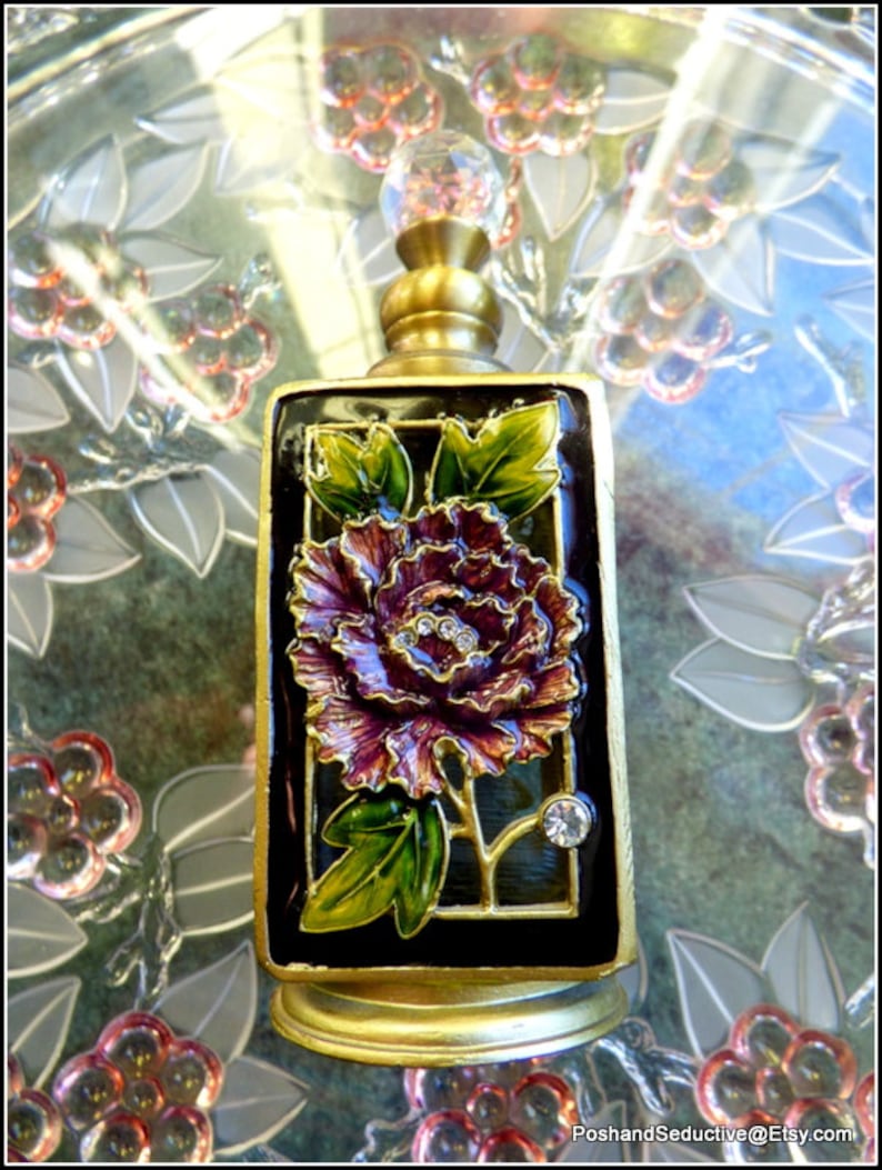 May include: A gold-toned perfume bottle with a black enamel frame and a floral design. The bottle features a large, purple flower with green leaves and a clear crystal accent.