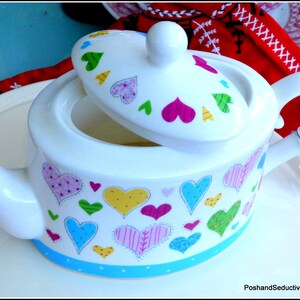 May include: White ceramic teapot with a lid decorated with colourful hearts. The teapot has a blue and white polka dot trim around the bottom.