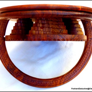 May include: A round wooden table with a unique design. The table top is made of multiple layers of wood, creating a tiered effect. The base of the table is a circular ring with intricate carvings.