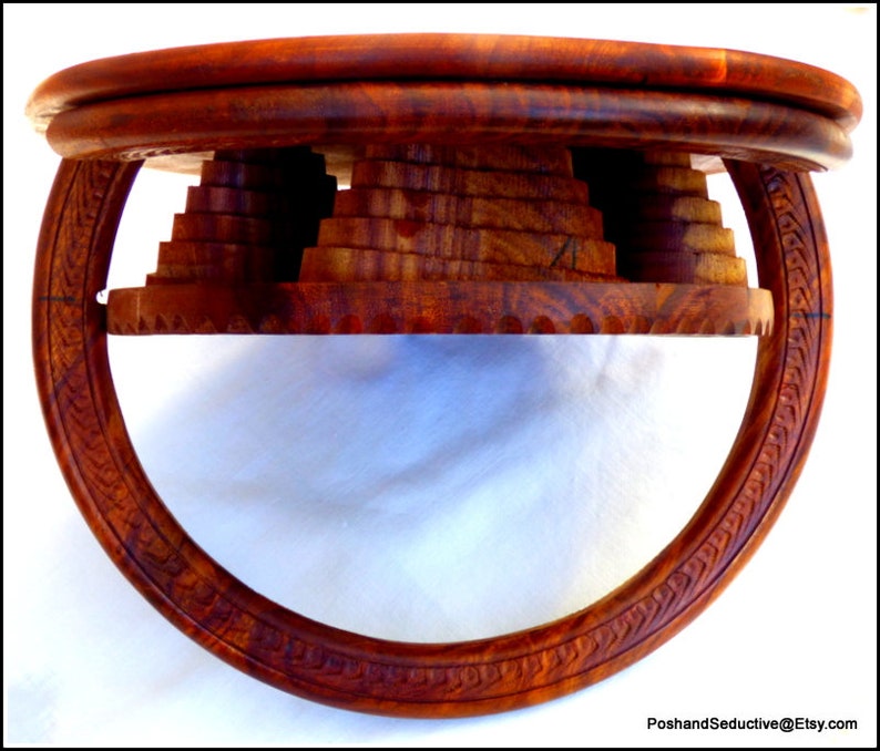 May include: A round wooden table with a unique design. The table top is made of multiple layers of wood, creating a tiered effect. The base of the table is a circular ring with intricate carvings.