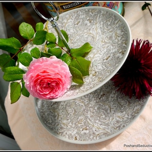Könnte beinhalten: Ein zweistöckiger Kuchenständer aus weißer Keramik mit goldenem Blumenmuster. Der Ständer ist mit einer rosa Rose und einer burgunderroten Dahlie verziert.