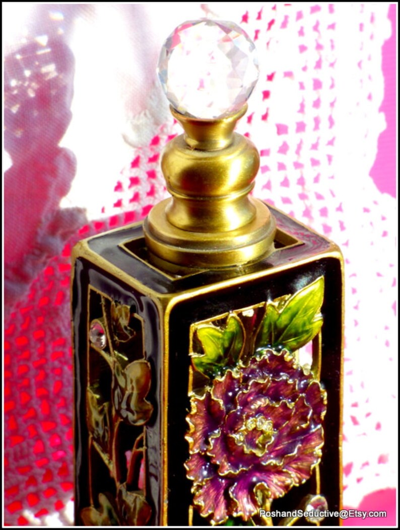 May include: A black and gold perfume bottle with a crystal stopper. The bottle is decorated with a purple flower and green leaves.
