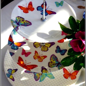 May include: Three-tiered white ceramic cake stand with a silver stand. Each tier features a different butterfly design with colourful wings. The bottom tier has a white background with small polka dots.