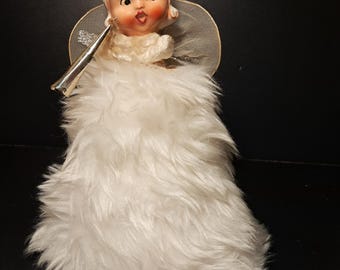 May include: A white porcelain angel tree topper with a white fur skirt and a silver trumpet. The angel has blonde hair, a red bow, and a painted face.