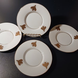 May include: Four white saucers with a gold bee design and gold trim. The saucers are arranged on a dark surface. The saucers have a circular indentation in the center. The bee design is repeated around the edge of each saucer.