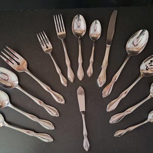 Puede incluir: Un conjunto de cubiertos pulidos, que incluyen tenedores, cucharas y cuchillos, dispuestos sobre una superficie oscura. La cubertería tiene mangos decorativos e incluye cucharas de servir y un cuchillo de mantequilla, ideal para una cena formal.