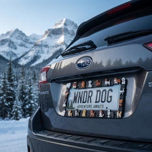 May include: A black license plate frame decorated with various small dog figurines attached to the border. The license plate inside features the text WNDR DOG and ADVENTURE AWAITS, along with a graphic of a mountain landscape and pine trees.