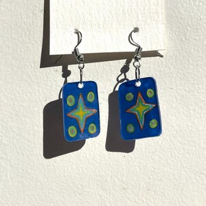 May include: A pair of blue rectangle-shaped earrings with a yellow star and green circles painted on them.