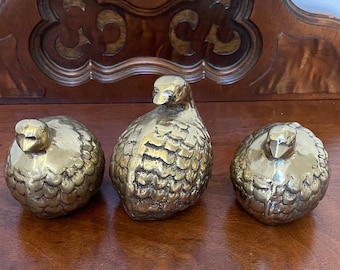 May include: Three gold-colored metal bird figurines. The birds are in a sitting position with their heads tilted down. The birds are on a wooden surface.