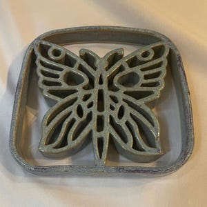 May include: A square ceramic trivet featuring a cutout butterfly design in the center. The piece has a glossy, mottled light blue and grey glaze with subtle purple undertones along the edges and within the intricate butterfly wing patterns.