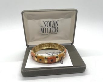 May include: A gold-toned hinged bracelet with orange and brown rectangular inlays, presented in a gray box. The bracelet features a decorative design and is from the "Nolan Miller Glamour Collection".