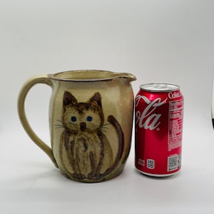 May include: A ceramic pitcher with a brown cat painted on the front. The pitcher is beige with brown accents and has a brown handle. The pitcher is next to a can of Coca-Cola.