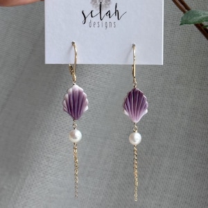 Puede incluir: Un par de pendientes de oro con cierre de palanca, con una cuenta morada en forma de concha, una perla blanca y una delicada cadena dorada colgante. Los pendientes se presentan en una tarjeta blanca con el texto Selah designs y un logo mandala.