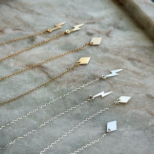 May include: Gold and silver chain earrings with lightning bolt and diamond-shaped charms. The earrings are displayed on a marble tray. The chains are delicate and dangle, showcasing the charms.